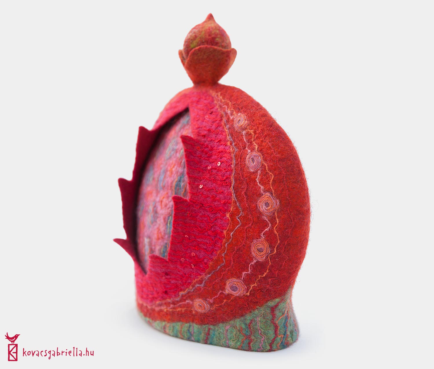 Tehuva i filtad ull med en knopp i form av en knopp. Röd och grön med intrikata inläggningar av trådar. Framsidan är filtad i två lager, där det övre lagret har klippts upp och avslöjar ett undre lager som är rutigt. Av Gabriella Kovács.