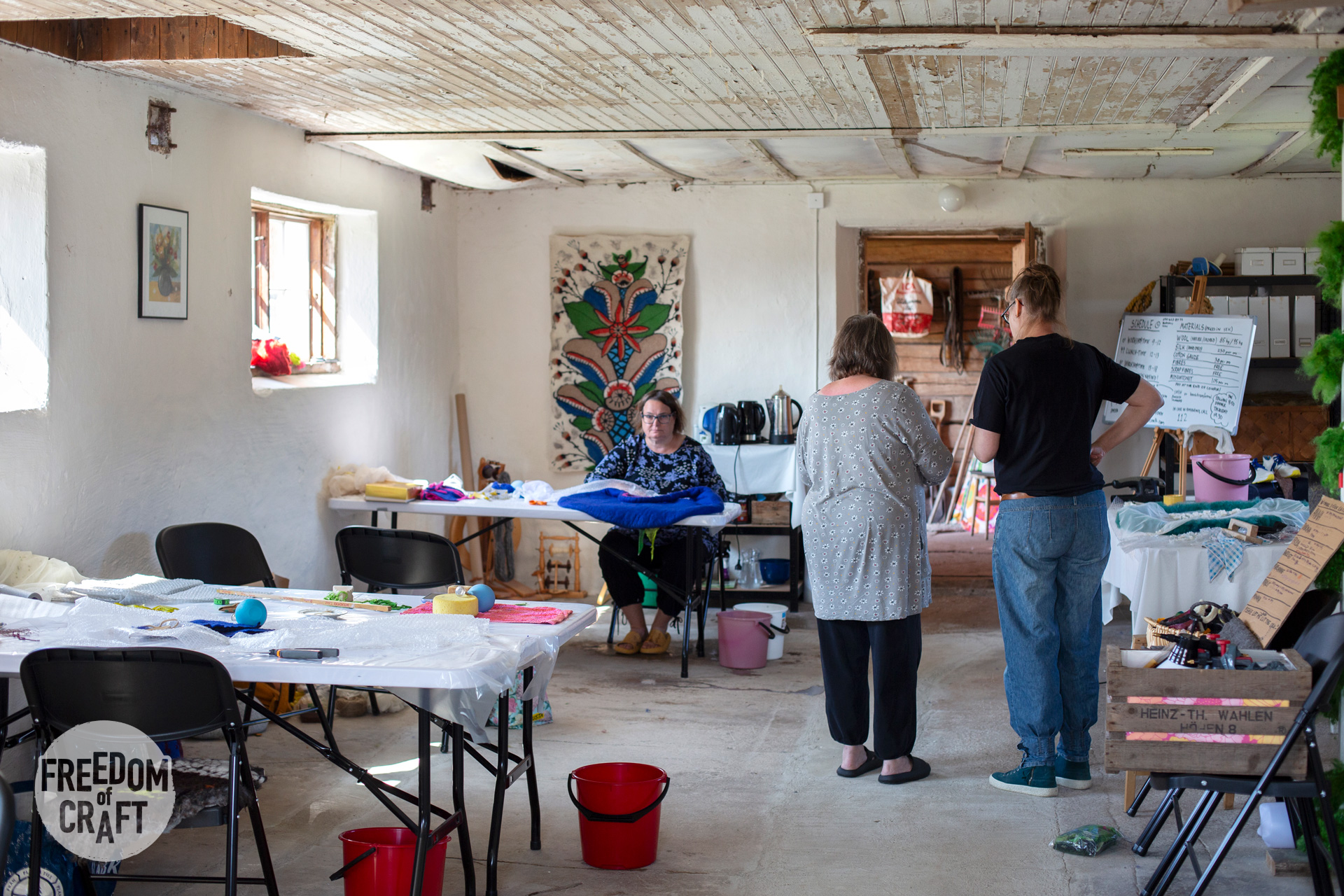A white room, rustic. Tables on the left, with ongoing felting projects. Two women at the far end is engaged in a discussion.