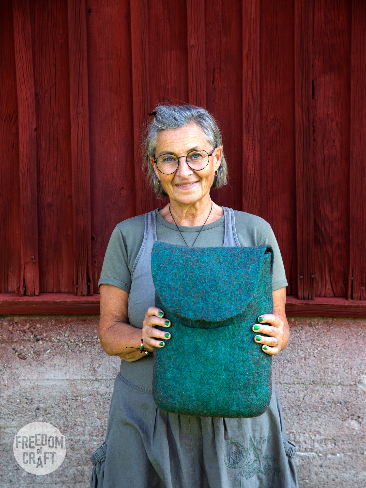 A woman holding up her back pack that she has made. It is turqoise-green and is matching her nail polish and eyes.