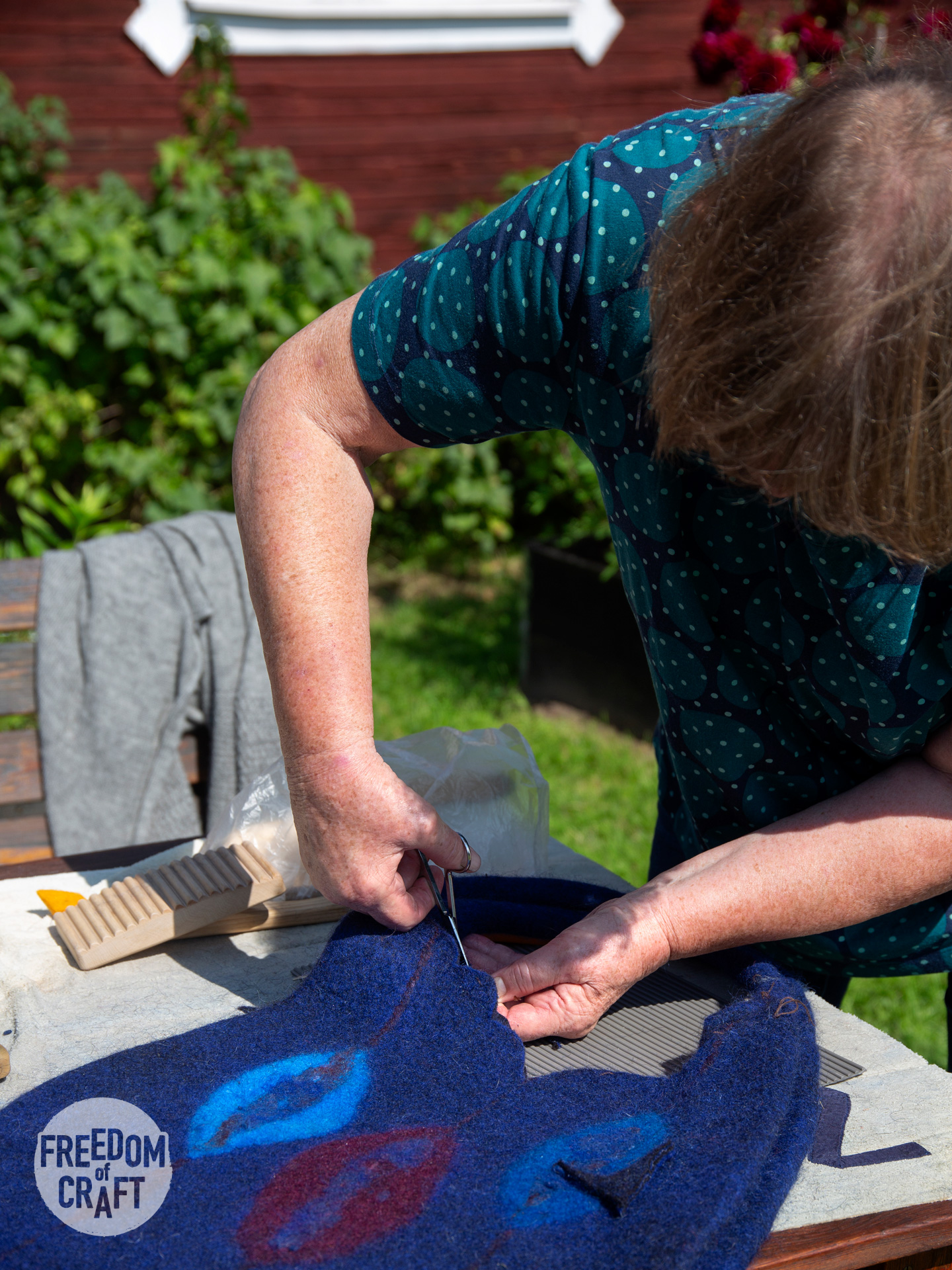 A woman is bent over her blue bag, cutting the edge of the opening outside in the summer sun.