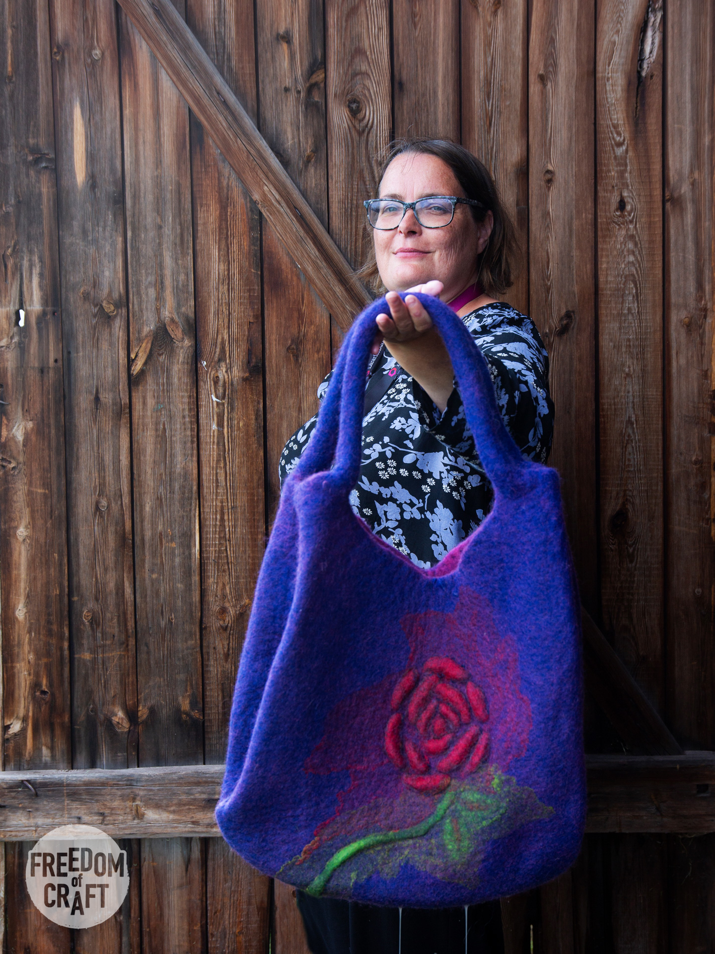 A woman with a proud expression is holding up her bag. It is lilac, with a big red rose in hot pink, silk and a green stem, with leaves.