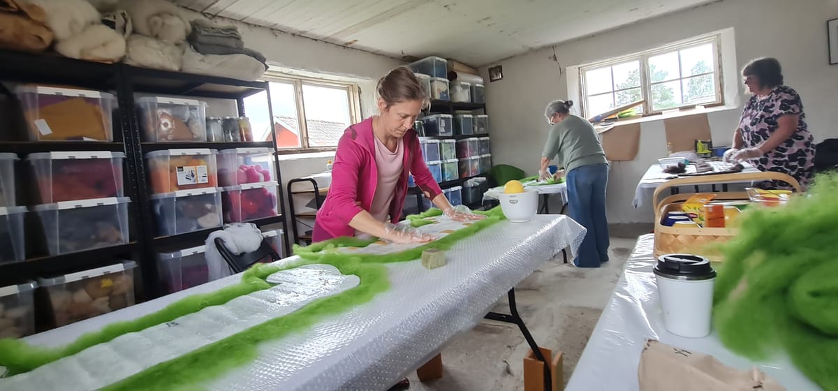 En kvinna i rosa arbetar med sitt skoprojekt. Grön ull ligger under en mall i plast.